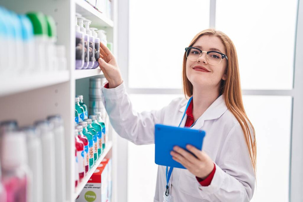Giovane donna rossa farmacista utilizzando touchpad che lavora in farmacia - Foto, immagini