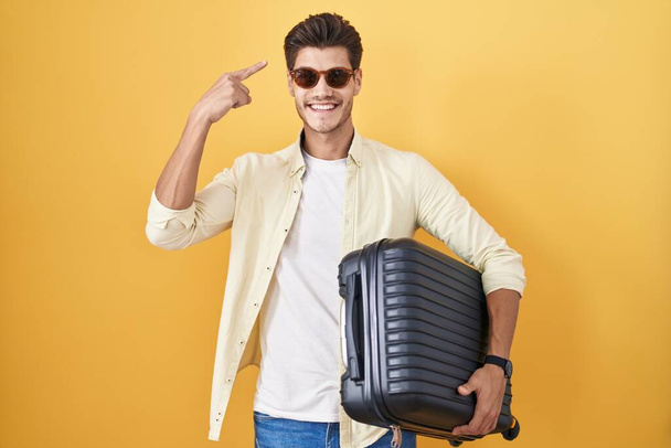 Молодий іспаномовний чоловік тримає валізу, що йде на літні канікули посміхаючись, вказуючи на голову одним пальцем, чудовою ідеєю або думкою, хорошою пам'яттю  - Фото, зображення