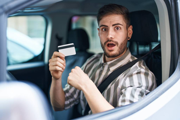 Latynoski mężczyzna z brodą prowadzi samochód trzymając kartę kredytową przerażony i zdumiony otwartymi ustami dla zaskoczenia, niedowierzanie twarz  - Zdjęcie, obraz