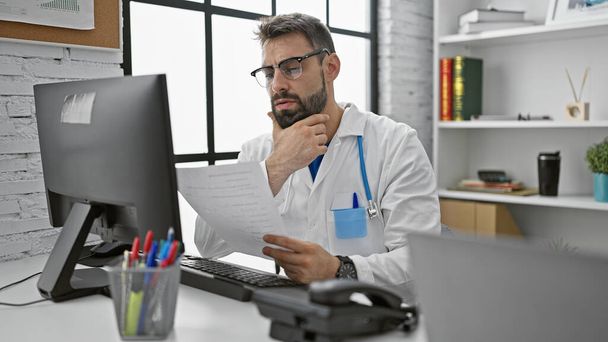 Junge und ernsthafte hispanische Arzt in seine Arbeit vertieft, Lesen medizinischer Bericht am Computer in der Klinik - Foto, Bild