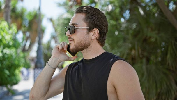 Junger hispanischer Mann mit Sonnenbrille spricht im Park mit Smartphone - Foto, Bild
