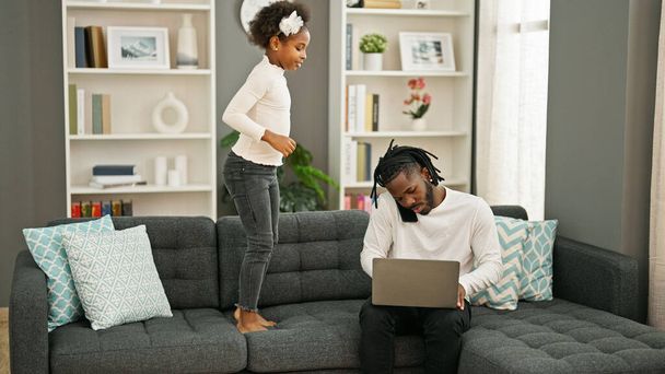 Le père et la fille afro-américains assis sur le canapé travaillent pendant que l'enfant dérange à la maison - Photo, image