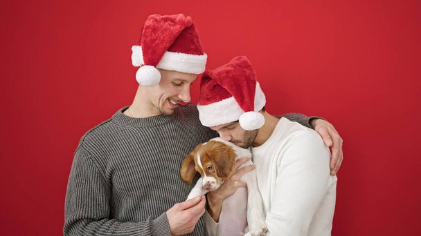 Twee mannen paar dragen kerst hoed zoenen hond over geïsoleerde rode achtergrond - Foto, afbeelding
