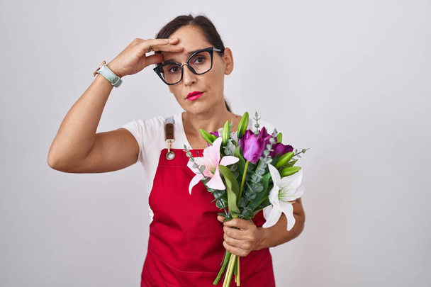Femme brune d'âge moyen portant un tablier travaillant dans un magasin de fleuristes tenant un bouquet inquiète et stressée par un problème avec la main sur le front, nerveuse et anxieuse de crise  - Photo, image