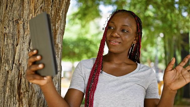 Fröhliche, selbstbewusste afrikanisch-amerikanische Frau genießt einen sonnigen Tag im Park, strahlt ein strahlendes Lächeln, während sie einen Online-Videoanruf auf ihrem Laptop tätigt - Foto, Bild