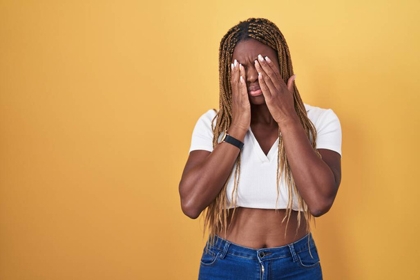 Afroameričanka s vlasy spletenými do copu, stojící nad žlutým pozadím, mnula si oči pro únavu a bolest hlavy, ospalý a unavený výraz. problém s viděním  - Fotografie, Obrázek