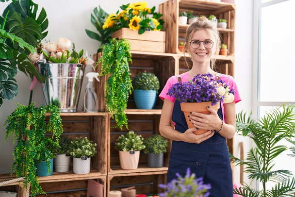Молодая блондинка-флористка улыбается уверенно держа растение в цветочном магазине - Фото, изображение