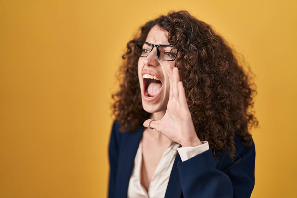 Ισπανίδα γυναίκα με σγουρά μαλλιά που στέκεται πάνω από κίτρινο φόντο φωνάζοντας και ουρλιάζοντας δυνατά στο πλάι με το χέρι στο στόμα. έννοια επικοινωνίας.  - Φωτογραφία, εικόνα