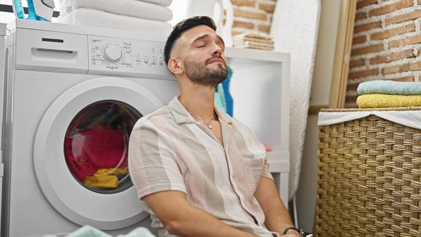 Young hispanic man leaning on washing machine stressed at laundry room - 写真・画像