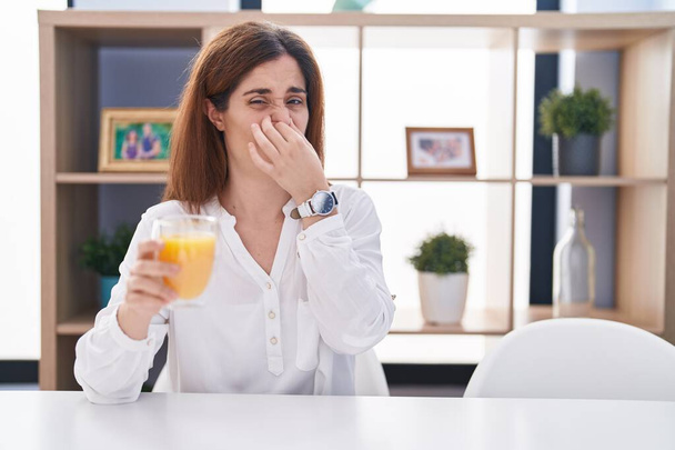 Brunette woman drinking glass of orange juice smelling something stinky and disgusting, intolerable smell, holding breath with fingers on nose. bad smell  - Photo, Image
