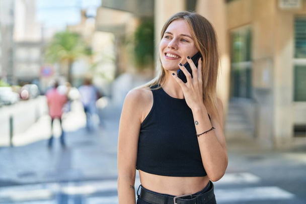 Junge blonde Frau lächelt selbstbewusst und spricht auf der Straße mit dem Smartphone - Foto, Bild