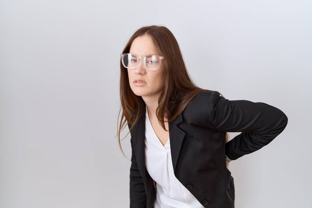 Belle femme brune portant une veste d'affaires et des lunettes souffrant de maux de dos, touchant le dos avec la main, douleurs musculaires  - Photo, image