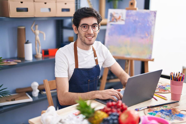 Молодий іспаномовний художник посміхається впевнено використовуючи ноутбук в художній студії
 - Фото, зображення
