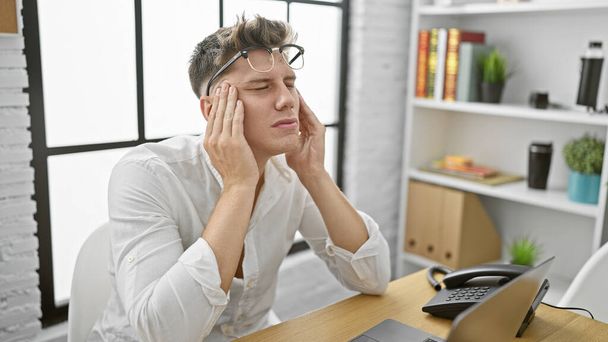 Zmartwiony młody biały mężczyzna cierpiący na ból głowy w pracy, zdjął okulary, siedział w biurze z laptopem - Zdjęcie, obraz