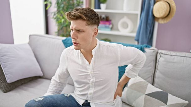 Endişeli beyaz bir adam ciddi bir sırt ağrısı, ızdırap içinde, oturma odasındaki kanepede oturuyor, arka planda, sırt ağrısından yaralı ve mutsuz. - Fotoğraf, Görsel