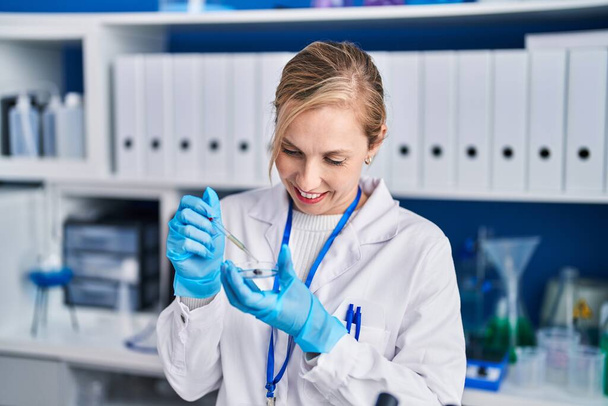 Junge blonde Wissenschaftlerin gießt Flüssigkeit auf Probe im Labor - Foto, Bild