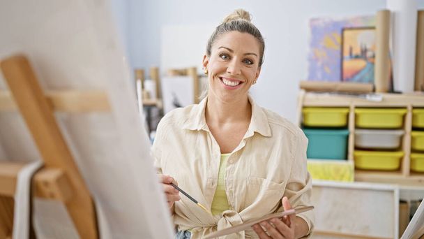 Junge blonde Künstlerin lächelt selbstbewusst und hält Pinsel und Palette im Kunstatelier - Foto, Bild