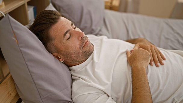 Retrato cautivador de un apuesto joven hispano agotado, profundamente relajante, dormido, descansando cómodamente, cómodamente acostado en la cama en el interior de su moderno dormitorio - Foto, imagen