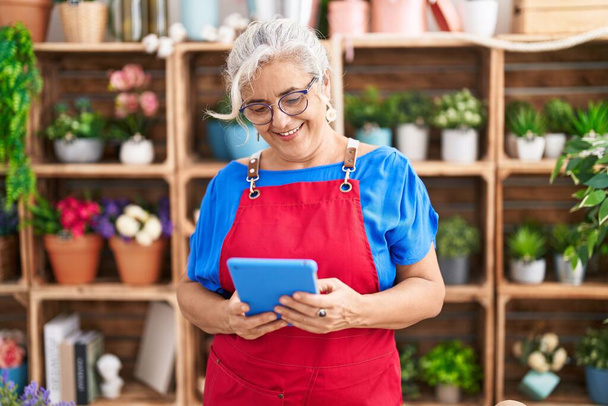 Florista mujer de pelo gris de mediana edad sonriendo confiada usando touchpad en floristería - Foto, Imagen