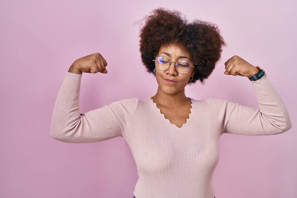 Joven mujer afroamericana de pie sobre fondo rosa mostrando los músculos de los brazos sonriendo orgulloso. concepto de fitness.  - Foto, Imagen