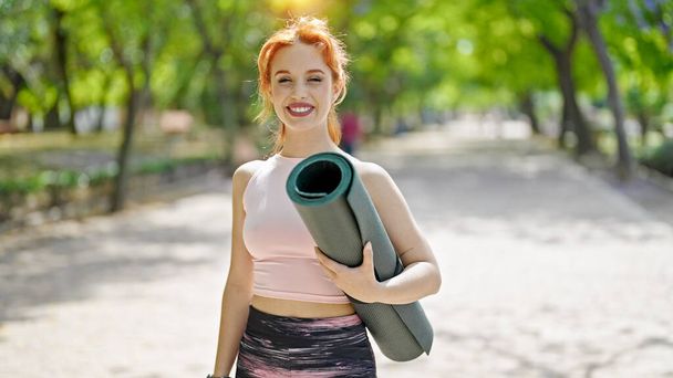 Молодая рыжая женщина, улыбающаяся в спортивной одежде, держит коврик для йоги в парке - Фото, изображение