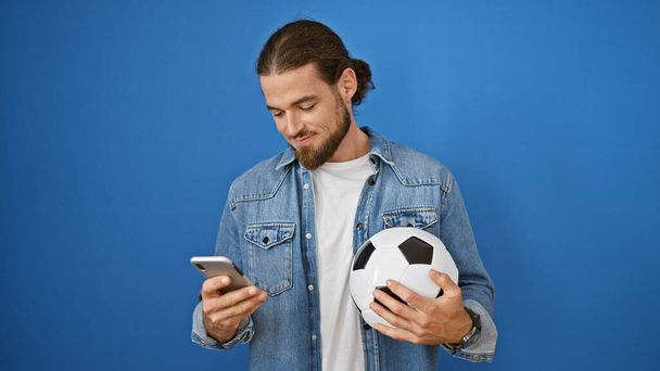 Giovane uomo ispanico sorridente tenendo palla da calcio e smartphone su sfondo blu isolato - Foto, immagini