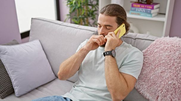 Un joven hispano hablando por teléfono sentado en el sofá bostezando en casa - Foto, imagen