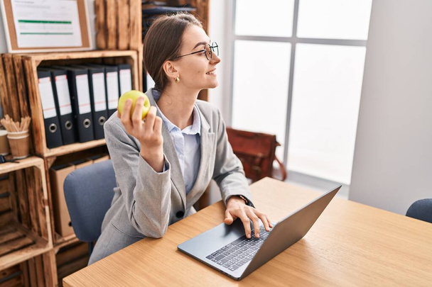Junge kaukasische Geschäftsfrau mit Laptop, der im Büro Apfel isst - Foto, Bild