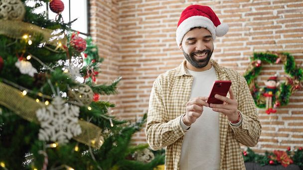 Joven hombre hispano celebrando la Navidad usando smartphone en casa - Foto, imagen