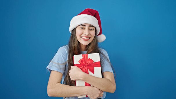 クリスマスの帽子をかぶった若い美しいヒスパニックの女性は孤立した青い背景に贈り物を抱きしめます - 写真・画像