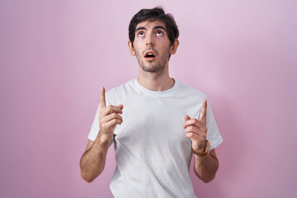 Young hispanic man standing over pink background amazed and surprised looking up and pointing with fingers and raised arms.  - Fotó, kép