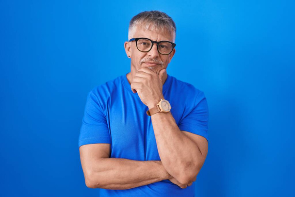 Hispanic man with grey hair standing over blue background with hand on chin thinking about question, pensive expression. smiling and thoughtful face. doubt concept.  - Photo, Image