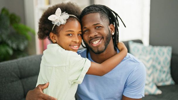 Afrikai amerikai apa és lánya magabiztosan mosolyogva ölelgetik egymást otthon, a kanapén ülve. - Fotó, kép