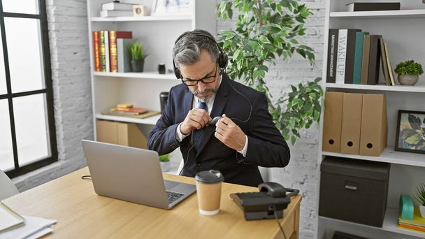 Attraktive grauhaarige junge hispanische Mann beherrscht das Büro Call Center Spiel, Video-Chat mit Client auf Laptop, geschmückt mit Anzug und Krawatte, hören coole Melodien. - Foto, Bild