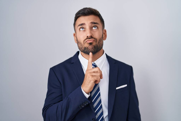 Pohledný hispánec v obleku a kravatě myšlení soustředěné na pochybnosti s prstem na bradě a vzhlíží zvědavý  - Fotografie, Obrázek