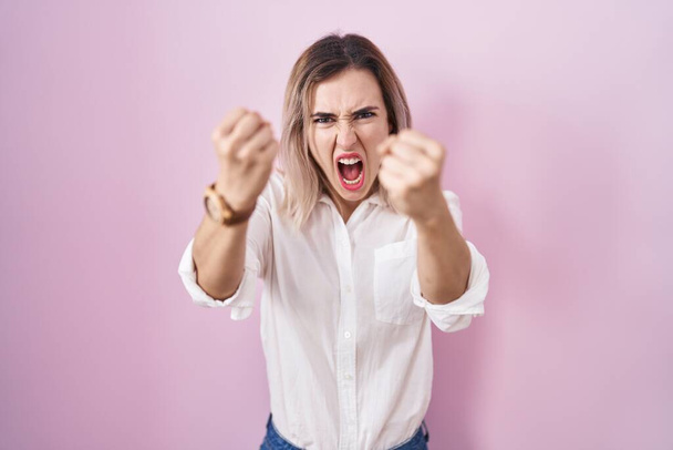 Молодая красивая женщина стоит на розовом фоне сердитый и сумасшедший поднимая кулаки разочарованы и в ярости, крича от гнева. гнев и агрессивная концепция.  - Фото, изображение