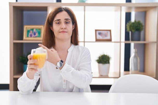 Bruna donna che beve bicchiere di succo d'arancia che punta con il dito della mano verso il lato mostrando pubblicità, viso serio e calmo  - Foto, immagini