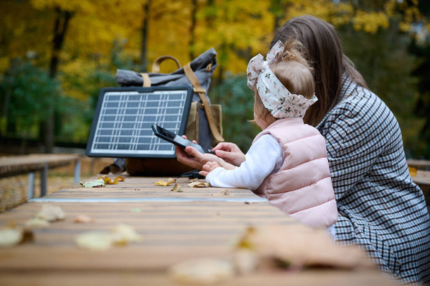Jeune fille mignonne avec sa mère marchant dans le parc avec des feuilles tombées, téléphone de charge avec panneau solaire portable. Concept d'énergie renouvelable. - Photo, image