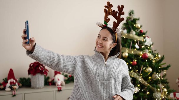 Young beautiful hispanic woman make selfie by smartphone standing by christmas tree at home - Foto, imagen