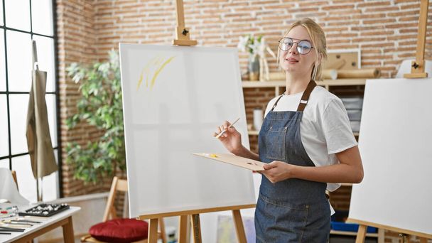 Jonge blonde vrouw artiest glimlachen zelfverzekerd holding penseel en palet in kunststudio - Foto, afbeelding