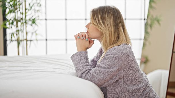 Young blonde woman leaning on bed praying at bedroom - 写真・画像