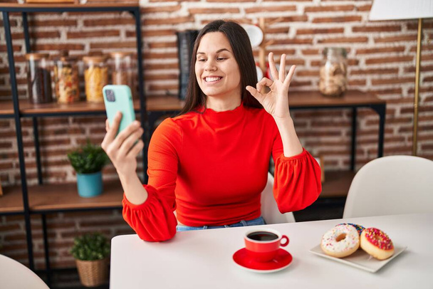 Nuori latino nainen syö donitseja ja juo kupin kahvia tekee videopuhelu tekee ok merkki sormilla, hymyillen ystävällinen ele erinomainen symboli  - Valokuva, kuva