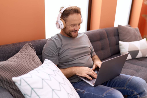 Hombre de mediana edad usando portátil y auriculares sentados en el sofá en casa - Foto, imagen
