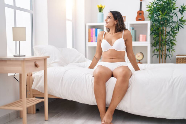 Молодая красивая латиноамериканка в нижнем белье сидит на кровати в спальне - Фото, изображение