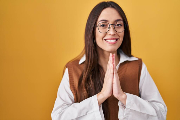Junge brünette Frau, die vor gelbem Hintergrund steht und eine Brille trägt und mit den Händen um Vergebung bittet, lächelt selbstbewusst.  - Foto, Bild