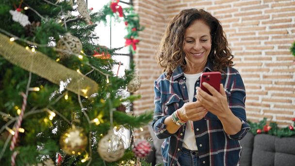 Mulher hispânica de meia idade usando smartphone celebrando o Natal em casa - Fotografia, imagem