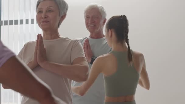 Mladá žena jóga učitel pomáhá veselý senior muž udržet rovnováhu v asana během skupinové praxe ve studiu - Záběry, video