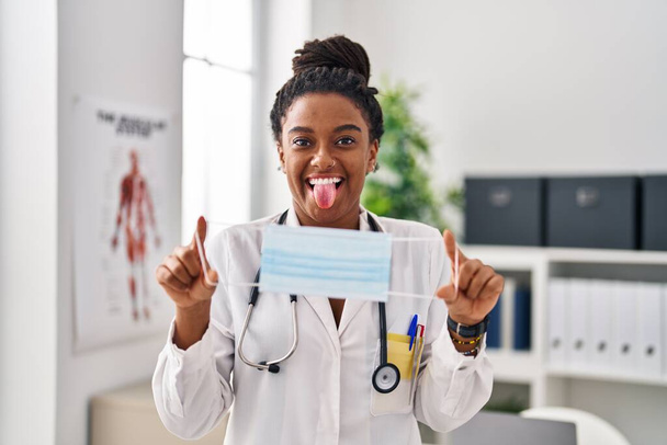 Young african american with braids wearing doctor uniform holding safety mask sticking tongue out happy with funny expression.  - Photo, Image