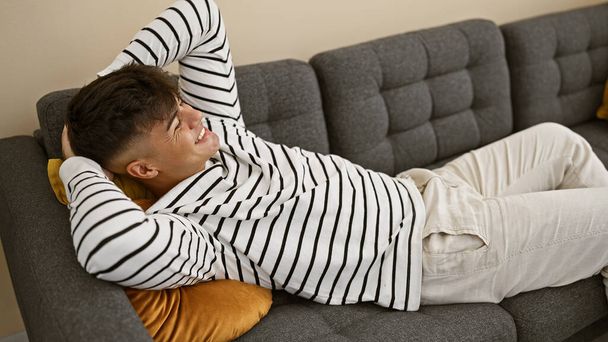 Junger, entspannter hispanischer Mann, bequem zu Hause auf einem gemütlichen Sofa liegend, die Hände hinter dem Kopf, verströmt im sonnendurchfluteten Wohnzimmer Zuversicht und Glück. - Foto, Bild