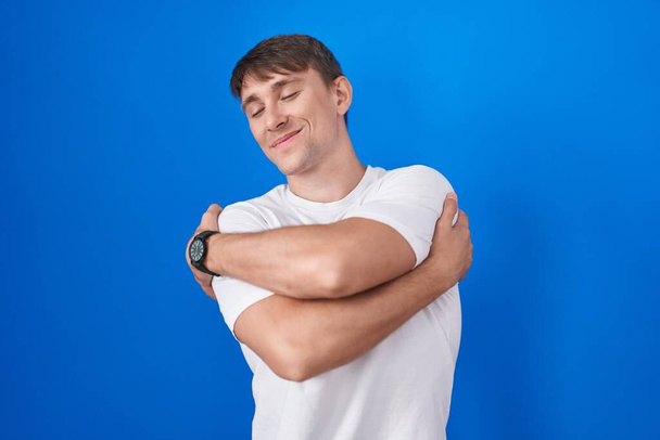Kaukasischer blonder Mann, der vor blauem Hintergrund steht und sich glücklich und positiv umarmt, selbstbewusst lächelt. Selbstliebe und Selbstfürsorge  - Foto, Bild